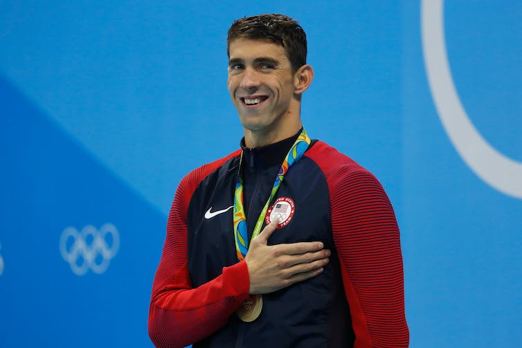 A smiling man wearing a gold medal around his neck holds his right hand over his heart.