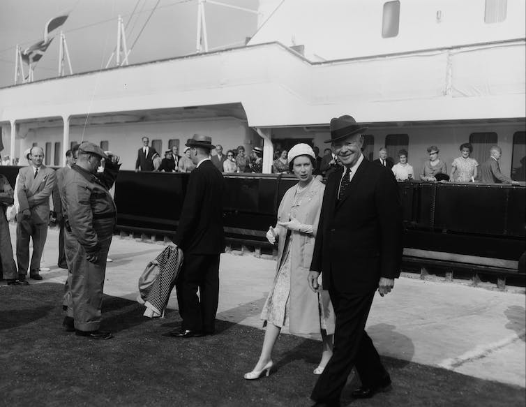 A black and white photo shows a woman in a pale-coloured suit with a man in a suit and a fedora.