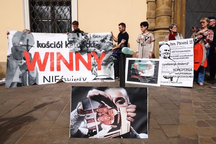 A handful of people hold red, white and black protest signs in front of a building, with a large photo of a woman ripping up a photograph in front of them.