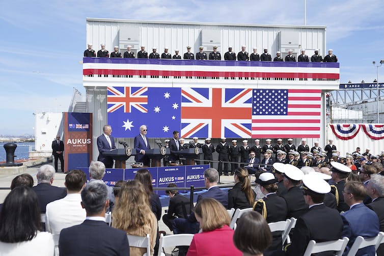 A crowd looks at a large sign with the Australian, U.K. and U.S. flags, while three men stand at lecterns just below it.
