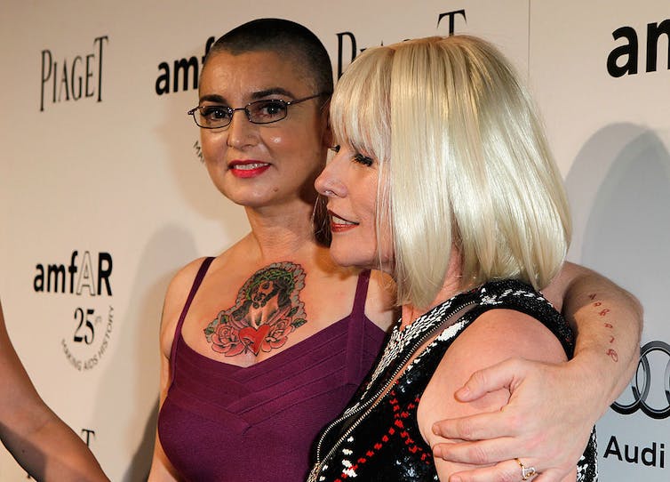 A woman in a purple dress with a shaved head and large, colorful tattoo stands embracing a blonde woman.
