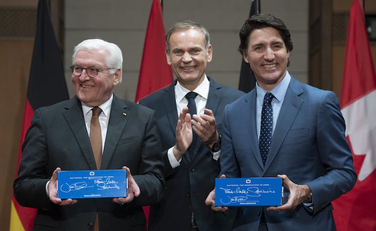 Three men, two holding batteries, smile for the cameras. A row of Canadian flags is behind them.