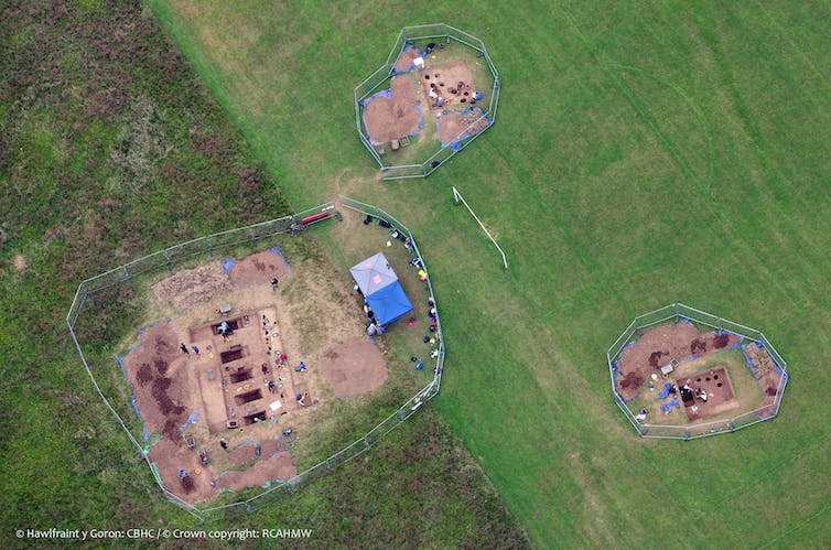 An aerial shot of a green field with three separate archaeological excavations taking place. There are precise holes in the ground and a blue tent set up.