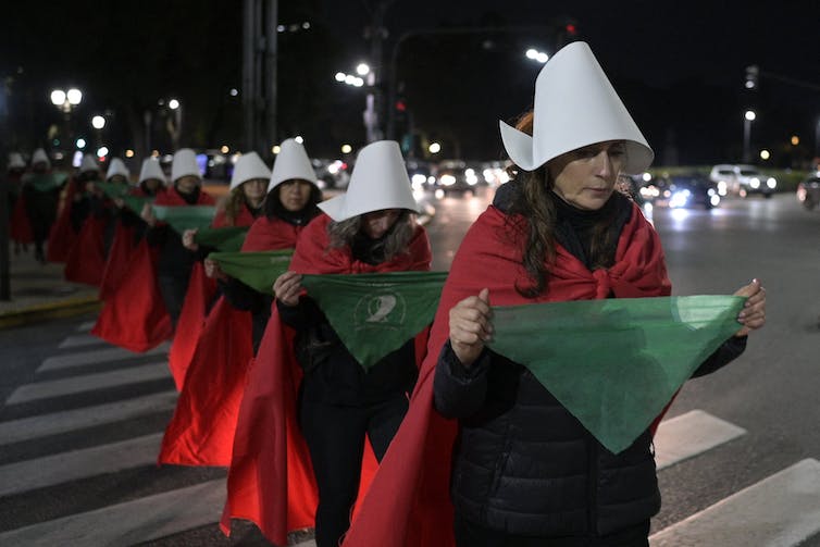 A row of women dressed like characters from the handmaids tale - wearing red robes and white hats - march in a line on a dark night.