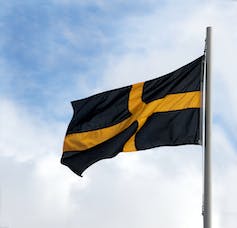 A black flag with a yellow cross flies with a blue sky in the background.