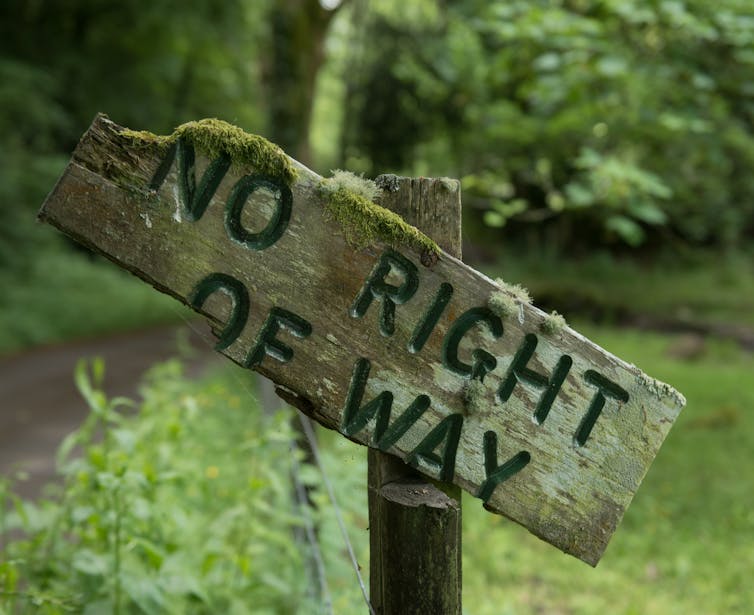 Close of up old broken wooden sign stating 'no right of way' with green leaves and countryside path in background